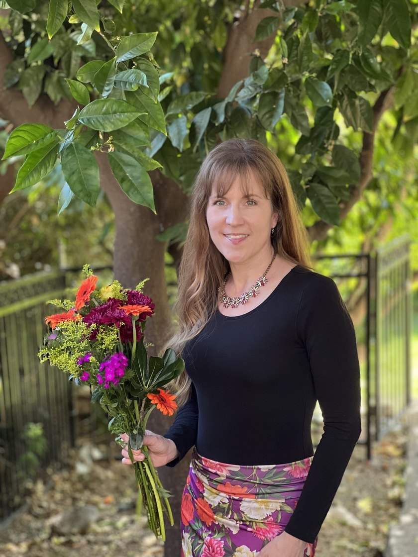 Heather Logghe holding a bouquet of wildflowers beneath a tree, in a black top and floral skirt