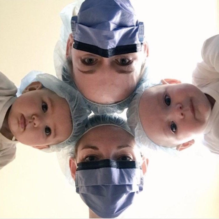 A surgeon in a cap and mask looking down into the camera with two babies in surgical caps beside her, a #ILookLikeASurgeon photo