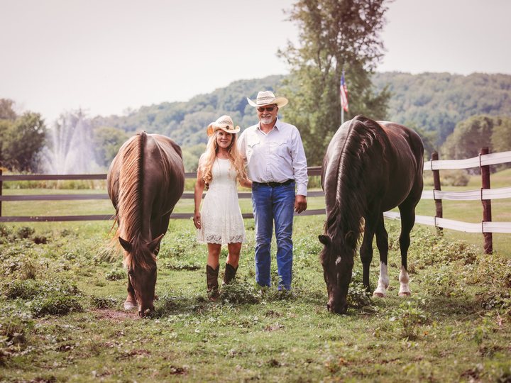 John and Jill Logghe standing between two horses in a pasture, wearing cowboy hats